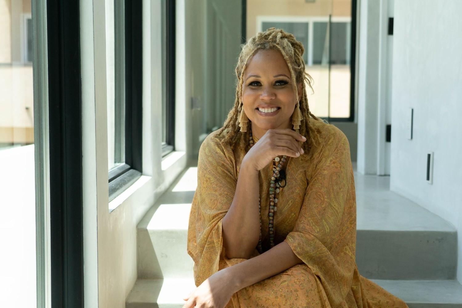 Breegan Jane sits in the hallway of her beach house. She is flanked by several large picture windows with black frames.