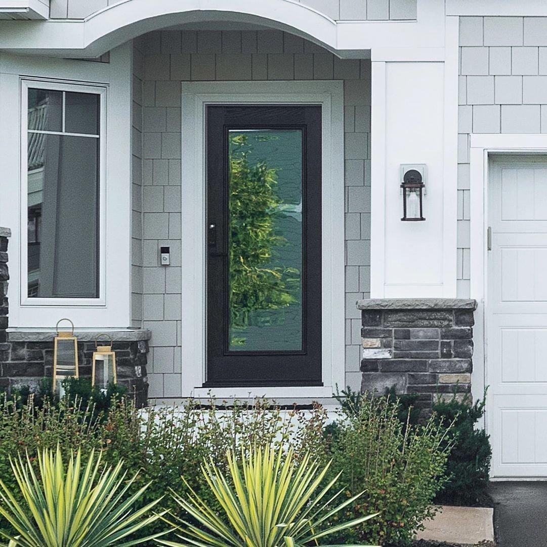 A single panel Ensemble entry door. The door is black and has full-light glass. It has white trim. You can see grey siding on the house and some spiky plants.