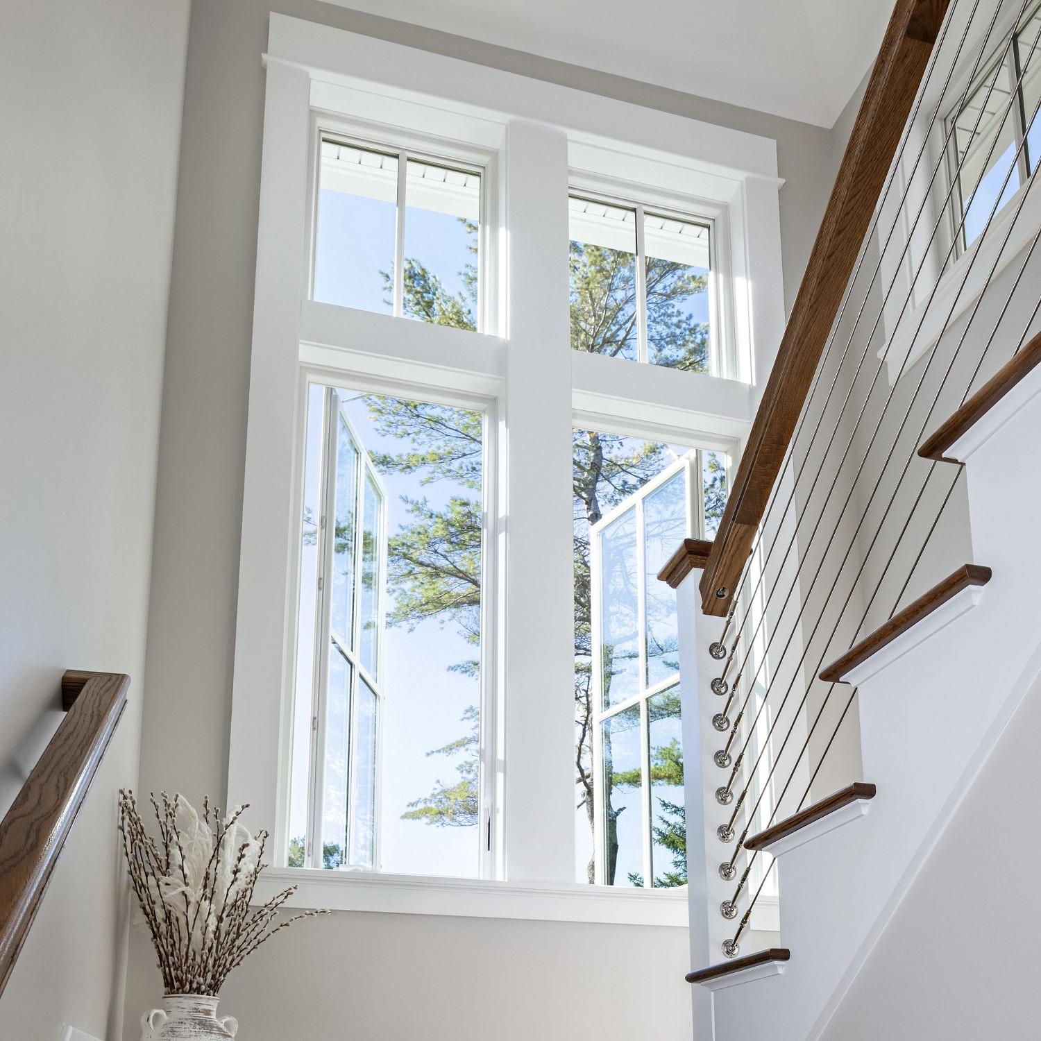 Looking upward into a stairwell with a set of large white casement windows topped with picture windows with grilles.