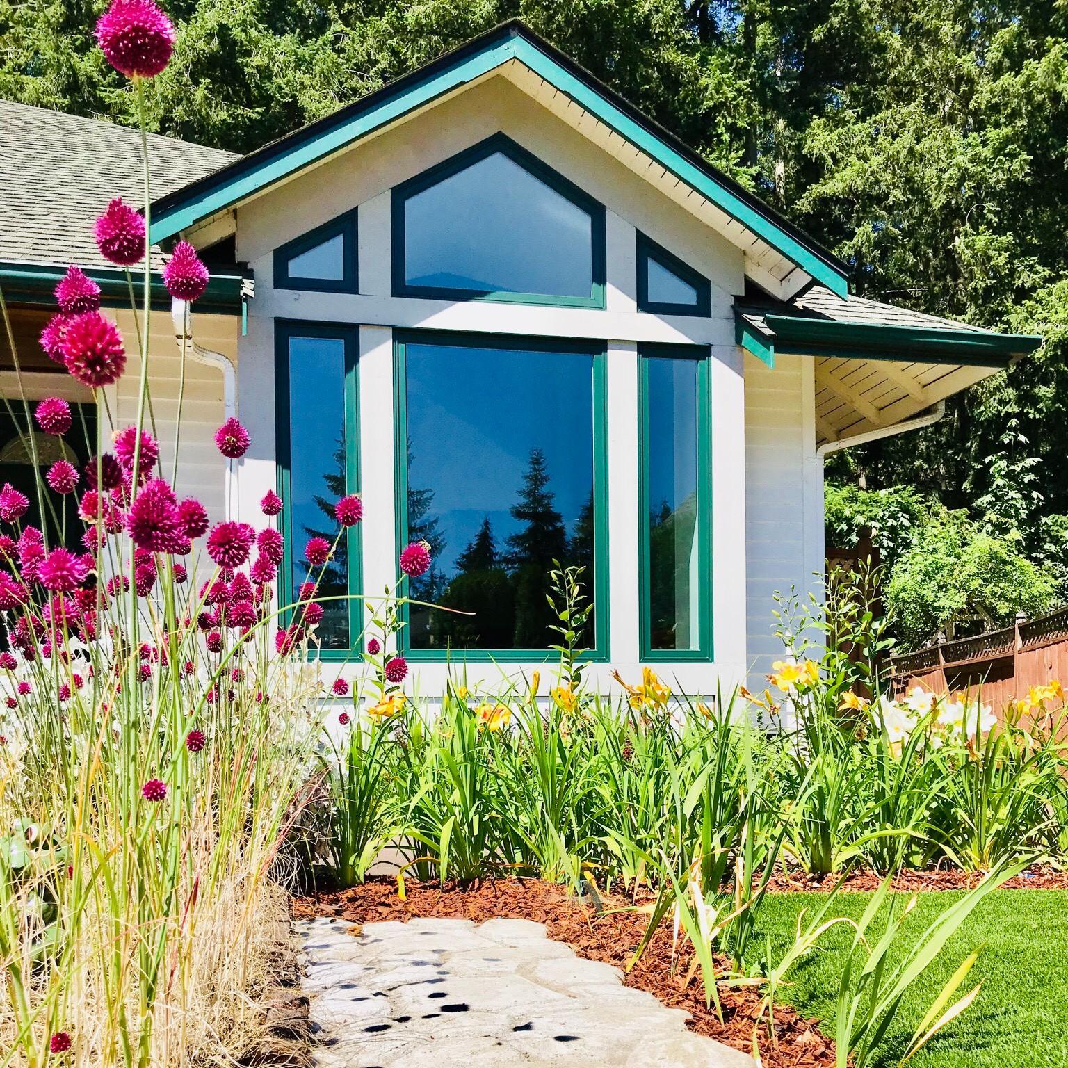 Two small trapezoid windows flank a peak pentagon window. These are set atop a bank of picture windows. The windows are forest green frames. There are pink flowers in the foreground providing contrast.