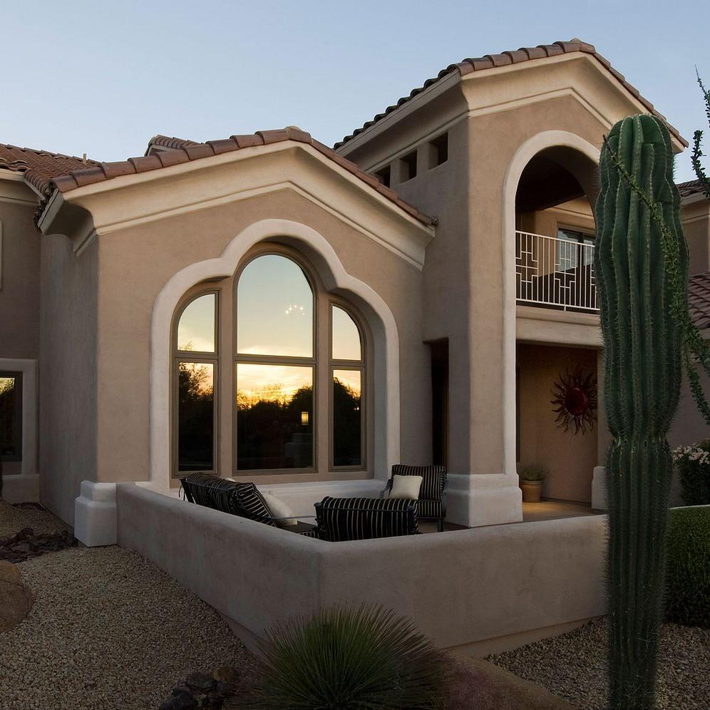 An adobe style home in the southwestern desert. The sunset is being reflected in the windows as twilight approaches. The home has picture windows topped with quarter circle and springline windows.
