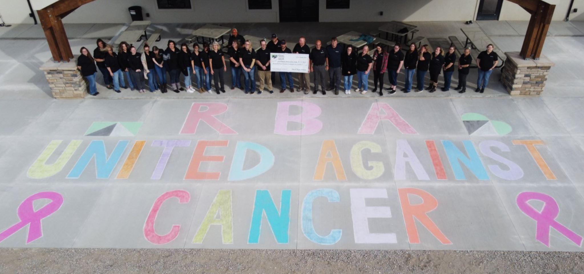 RbA United Against Cancer written on chalk in front of the team from Wyoming. They are holding a large check for a donation.